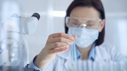 Scientist female researcher with protective glasses and mask examining lab tube with a blue fluid. Microbiology and science concept