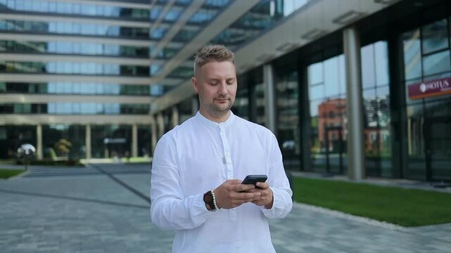 Focused young corporate worker investor businessman walking and using mobile phone outdoors near a business centre. High quality FullHD footage