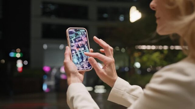 Woman holding smartphone with both hands. Phone screen showing map or navigation app with streets and buildings illuminated in bright colours. Woman wearing light jacket. Exploring map of night street