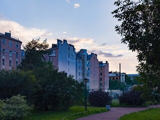 A serene evening scene featuring residential apartment buildings bathed in the soft light of dusk. The image captures a peaceful urban park with lush greenery in the foreground, contrasted against the