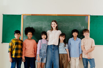 Diverse group of elementary students pose with teacher in front of classroom chalkboard. Multicultural children and educator showcase inclusive learning environment in primary school setting