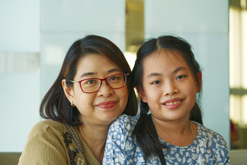 Portrait Asian mother and daughter sitting together, healthy middle- aged woman and child at 9 to 10 years old, both looking at camera, indoors image with natural light.