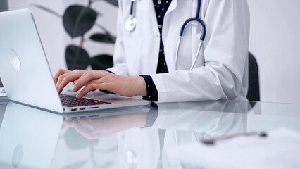Doctor woman is typing on laptop computer at glass table in clinic, horizontal view. Medicine and pharmacy concept