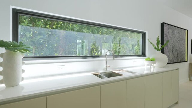 Unusual working area in sparse kitchen with oblong window above sink and tap installed in cabinets. Live plants in white vases serving as elements of decoration