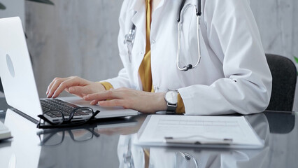 Doctor woman wearing white medical coat over a yellow blouse is working on laptop with stethoscope on the glass desk. Medicine and Health care