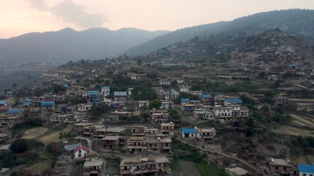 "Aerial view of a traditional village in Jumla, Nepal, nestled in the hills. The village features houses with blue roofs scattered across terraced fields, surrounded by rugged terrain and lush forests