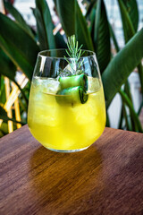 Stemless tulip glass filled with ice and a vibrant yellow cocktail, garnished with a fresh cucumber slice and a sprig of rosemary. Fresh, herbal touch