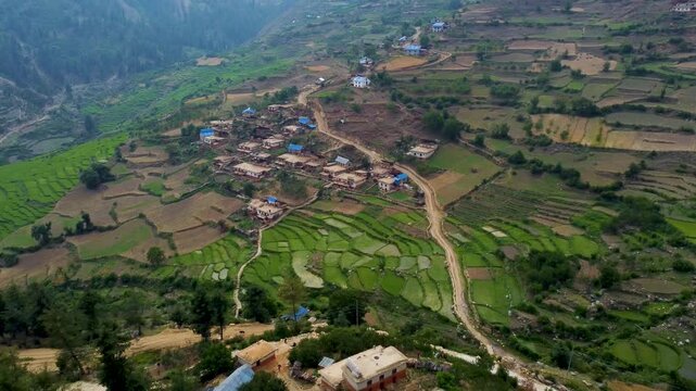 "Aerial view of a traditional village in Jumla, Nepal, nestled in the hills. The village features houses with blue roofs scattered across terraced fields, surrounded by rugged terrain and lush forests