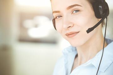 Beautiful female specialist with headphones helping customers online and smiling charmingly in sunny call center office. Business people concept