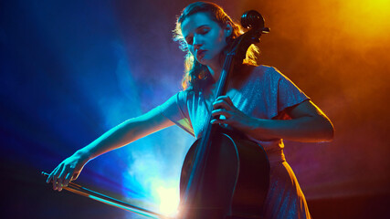 Young talented woman, musician, performing solo cello on stage surrounded dramatic smoke and neon light. Orchestra. Concept of classical and modern music, entertainment, jazz. Ad © master1305