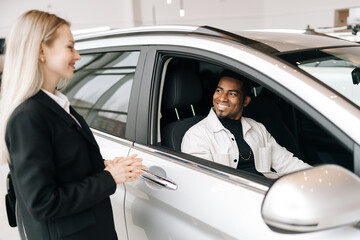 Cheerful African-American customer sitting behind steering wheel of new car in auto dealership, listening female consultant. Friendly auto dealer in business suit talking with black male customer.