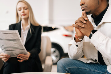 Low-angle view of female manager consulting doubtful black buyer, giving purchase agreement for review at showroom. Pensive African client receiving car purchase contract before signing in dealership