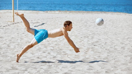 Dynamic game. Young man in blue shorts energetically playing beach volleyball, falling down on sand while hitting ball. Concept of sport, summer activity, leisure games, friendship © master1305