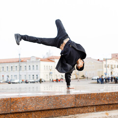 Cool fashionable man dancer with sunglasses in trendy fashion black clothes with cap, jacket, jeans and sneakers dancing on hand in city, Hipster guy in motion outdoors
