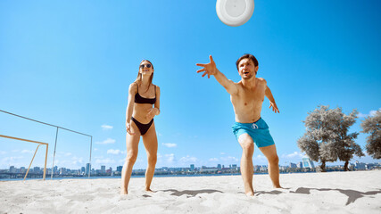 Young man and woman, friends playing dynamic frisbee game on beach by the river under clear blue...