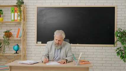 Aged man college professor sit at desk in classroom in front of chalkboard grading test papers, highlighting mistakes with red marker. Education concept.