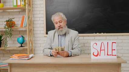Aged man college professor sit at desk in classroom in front of chalkboard with paper sign word sale on it and counting money. Education concept.