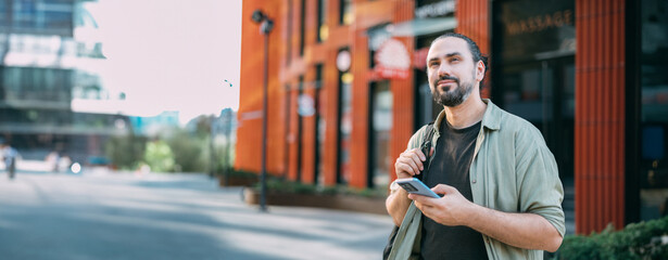 A young Caucasian man with a phone in his hands on a bright city street.