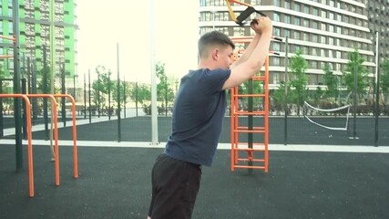 A teenager on an outdoor sports ground does arm exercises using TRX.