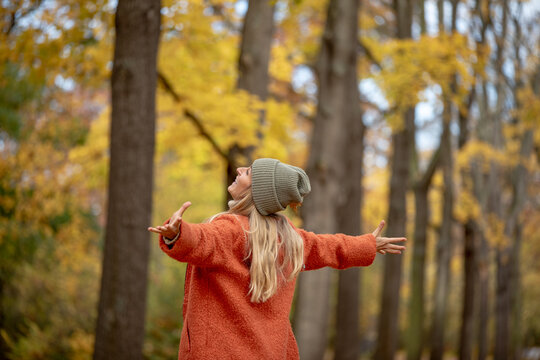 Person in an orange coat and gray beanie stands in a forest with arms outstretched, embracing the autumn season, surrounded by trees with golden leaves, exuding joy and freedom.