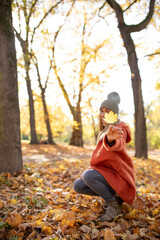 Woman kneels on the ground in an autumn forest, extending a yellow leaf towards the camera, wearing a cozy orange coat and knit hat, surrounded by fallen leaves and golden light
