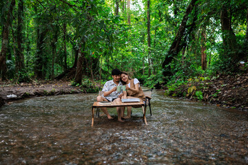 relax and romantic time concept, young asian couple enjoying sitting on wooden chairs drinking coffee together in middle of a stream