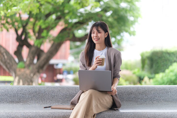 Businesswoman sitting on an outdoor bench, holding a coffee cup in one hand and her laptop on her lap.