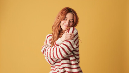 Cute red-haired woman dressed in sweater hugs herself looking at camera on yellow background....