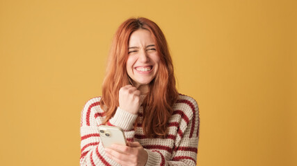 Red-haired girl dressed in sweater, with mobile phone in hands celebrates victory on yellow background. Beautiful girl poses in studio with blank space on yellow wall for your promotion