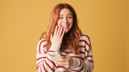 Surprised girl looking at camera while holding smartphone on yellow background