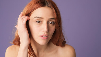 Close-up of red-haired woman looking at mirror in studio on purple background, POV shot. Beautiful girl poses in studio with blank space on violet wall for your promotion