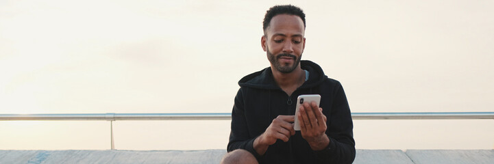 Close up, young smiling bearded male athlete dressed in hoodie uses cellphone after training while sitting on embankment, panorama