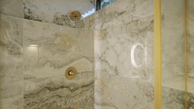 Marble tile in beige and grey colours on walls of shower cabin with tiny window and bronze plumbing. Propert interior in clean bathroom with reflected lightning