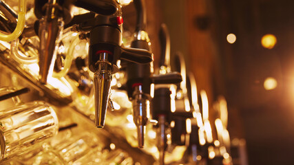 Close-up of multiple shiny beer taps with handles, set against blurred background with rows of empty glasses hanging from it. Cozy lights. Concept of beer, brewery, pub, Oktoberfest, traditions