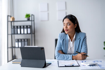 A woman in a blue suit sits at a desk with a laptop and a pen