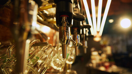 Close-up of multiple shiny beer taps with handles and mirrors, set against blurred background with rows of empty glasses and bar counter in the background. Concept of beer, brewery, pub, Oktoberfest