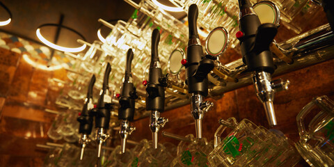 Row of shiny beer taps with handles, set against wall with rows of empty mugs hanging from it. Vintage and luxury atmosphere. Concept of beer, brewery, pub, Oktoberfest, traditions