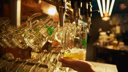 Bartender hand holding glass of lager beer being poured from tap, with rows of empty glasses hanging above and behind the tap. Concept of beer, brewery, pub, Oktoberfest, traditions