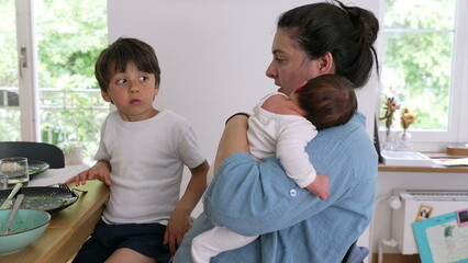 Obraz premium Mother holding baby while sitting next to an older child eating at the dining table. The scene reflects the multitasking and nurturing aspects of parenthood in a cozy home environment