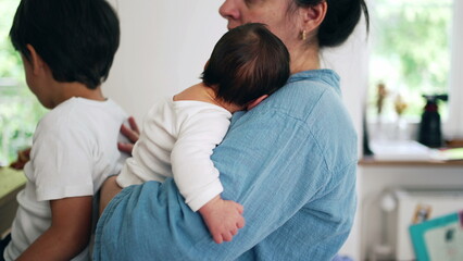 Mother holding newborn baby close while another child stands nearby eating. This image captures the...