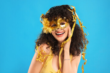 Happy young woman wearing carnival mask and tinsel on light blue background