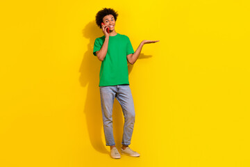 Full length photo of handsome good mood guy wear green t-shirt listening modern device showing arm emtpy space isolated yellow color background