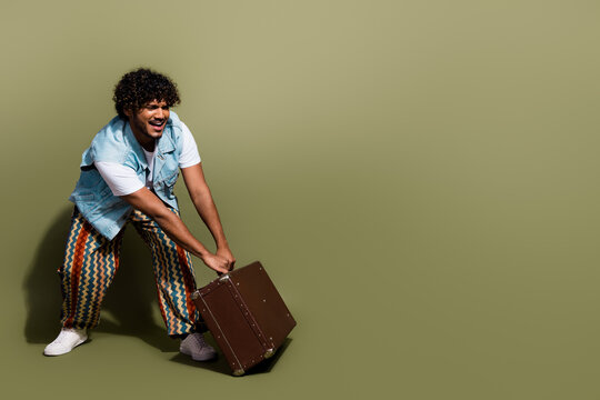 Full size photo of handsome young guy pull heavy retro valise wear trendy jeans outfit isolated on khaki color background
