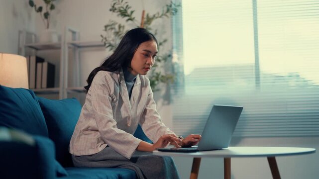 Young asian woman sits comfortably on a couch, focused on her laptop while working from home. The cozy living room setting provides a modern backdrop for her professional endeavors