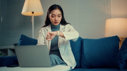 Young woman in casual business attire is sitting on a sofa, working on a laptop and checking her watch, potentially concerned about deadlines or upcoming appointments