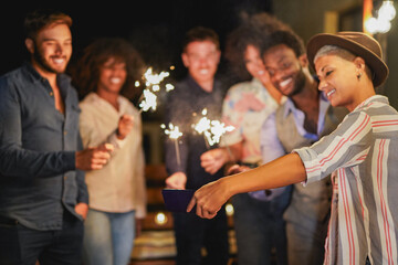 Group of multiracial friends taking a selfie during new year's eve at house terrace using firework sparklers - Soft focus on the hand holding mobile phone