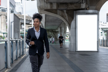 Fototapeta premium Teenage businesswoman, american african, afro hairstyle Holding a tablet walking on city street rushing to work. Empty white billboard for advertising.