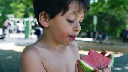 Young boy focused on eating a juicy watermelon slice, enjoying a sunny day in the park, shirtless with a relaxed expression, healthy summer snack, refreshing and hydrating, outdoor fun