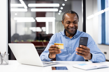 African american mature businessman holding credit card while using smartphone for online transaction inside contemporary office.
