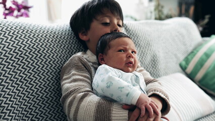 Young boy in a cozy sweater cradling his newborn sibling on a patterned sofa. baby rests peacefully while the boy holds them securely, serene family moment of bonding